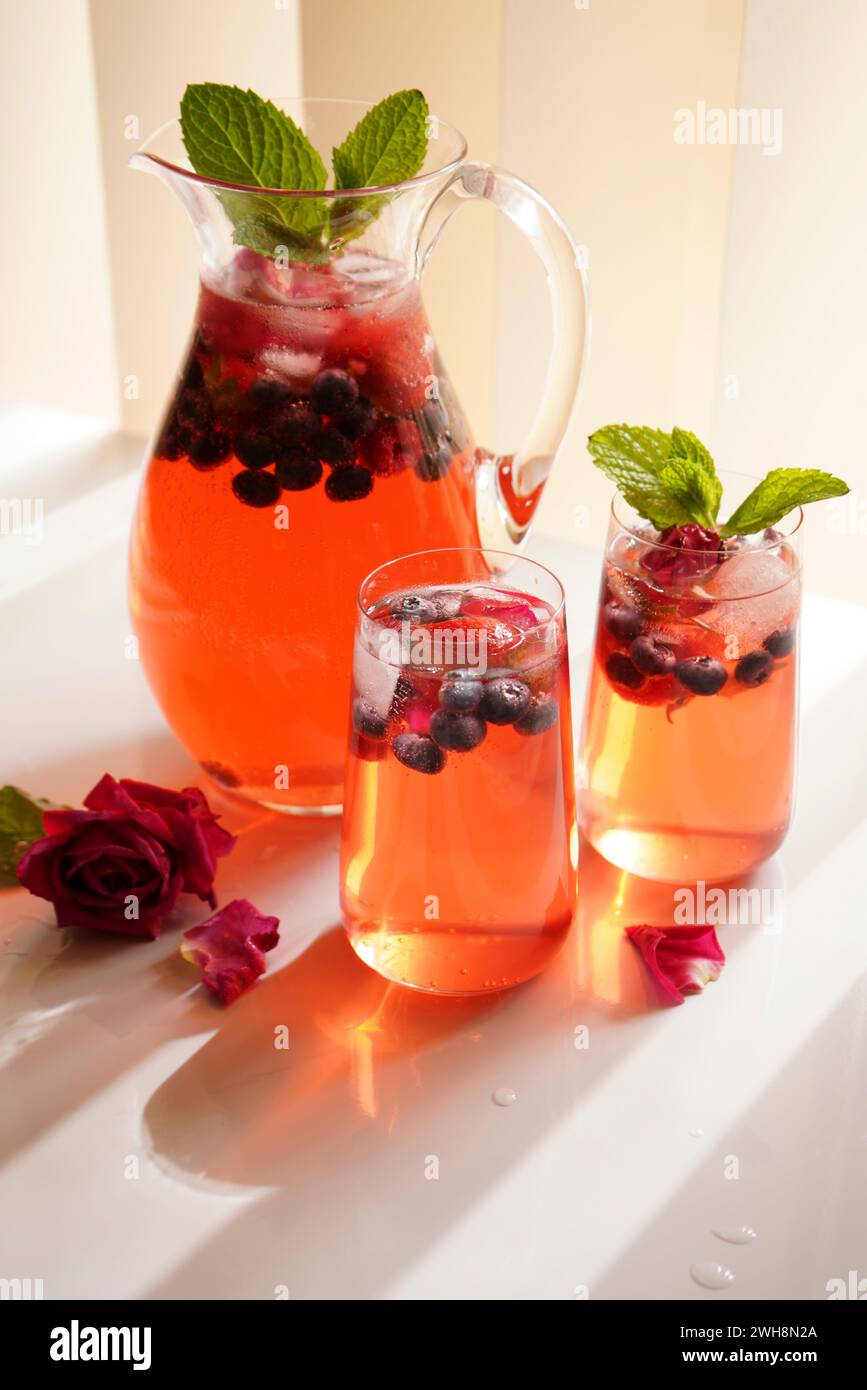 Spritzer di frutti di bosco e petali di rosa rinfrescante bevanda estiva sul tavolo bianco. Primo piano della brocca e due bicchieri. Foto Stock