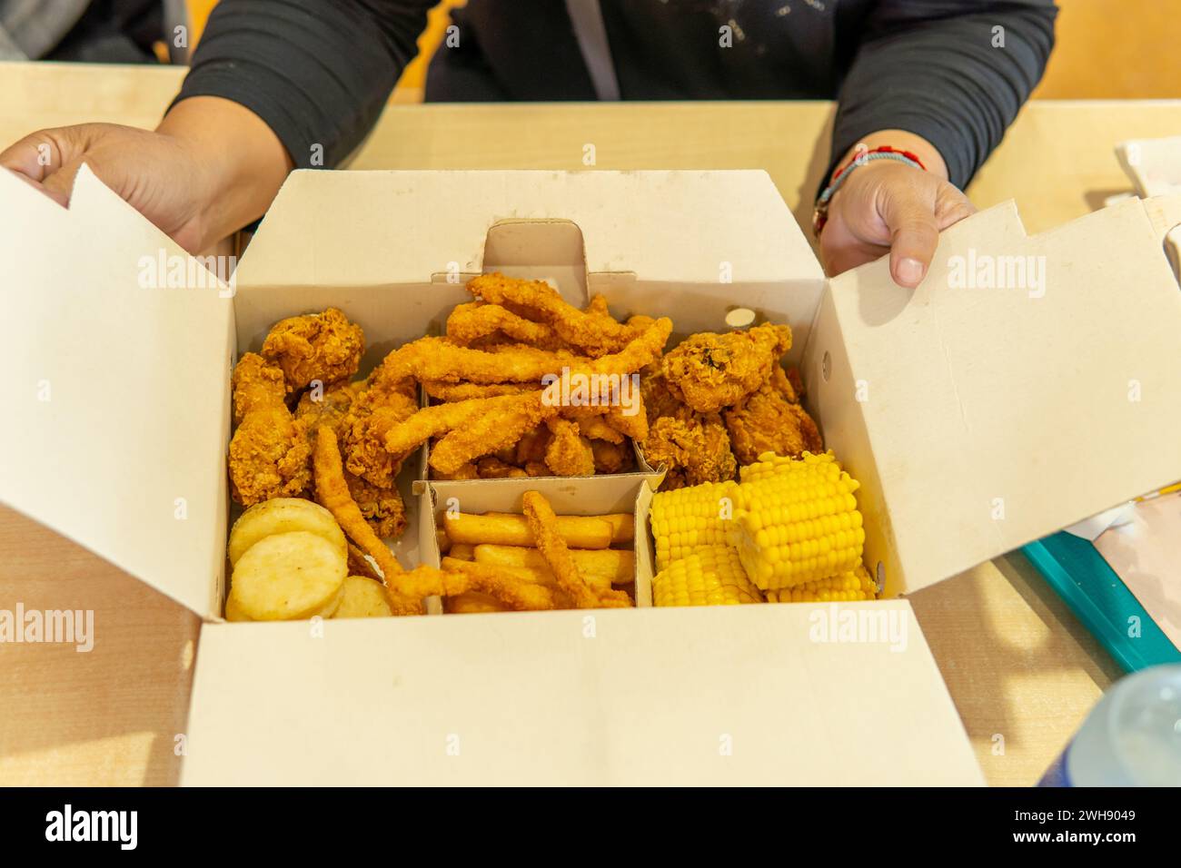 mani di una persona irriconoscibile che apre una scatola di fast food, con pollo, mais e patatine fritte su un tavolo Foto Stock