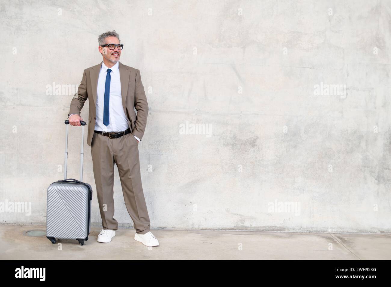 Un uomo d'affari maturo in una tuta intelligente si regge con una valigia, con un sorriso allegro su uno sfondo di cemento testurizzato, che racchiude la facilità di un moderno viaggio d'affari. Foto Stock