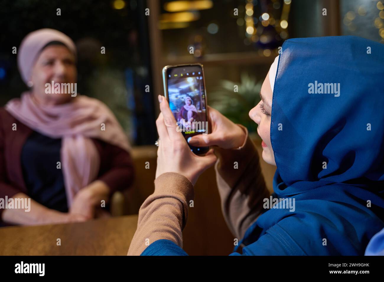 In un ristorante moderno durante il mese sacro del Ramadan, una donna in un hijab cattura un momento con sua madre utilizzando uno smartphone, simboleggiando il Foto Stock