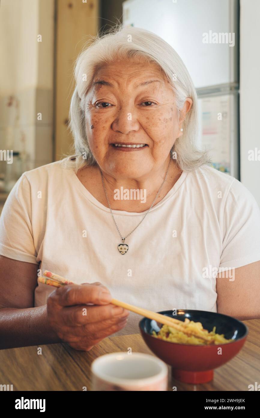 Anziana donna asiatica che mangia riso con bacchette guarda e sorride alla macchina fotografica Foto Stock