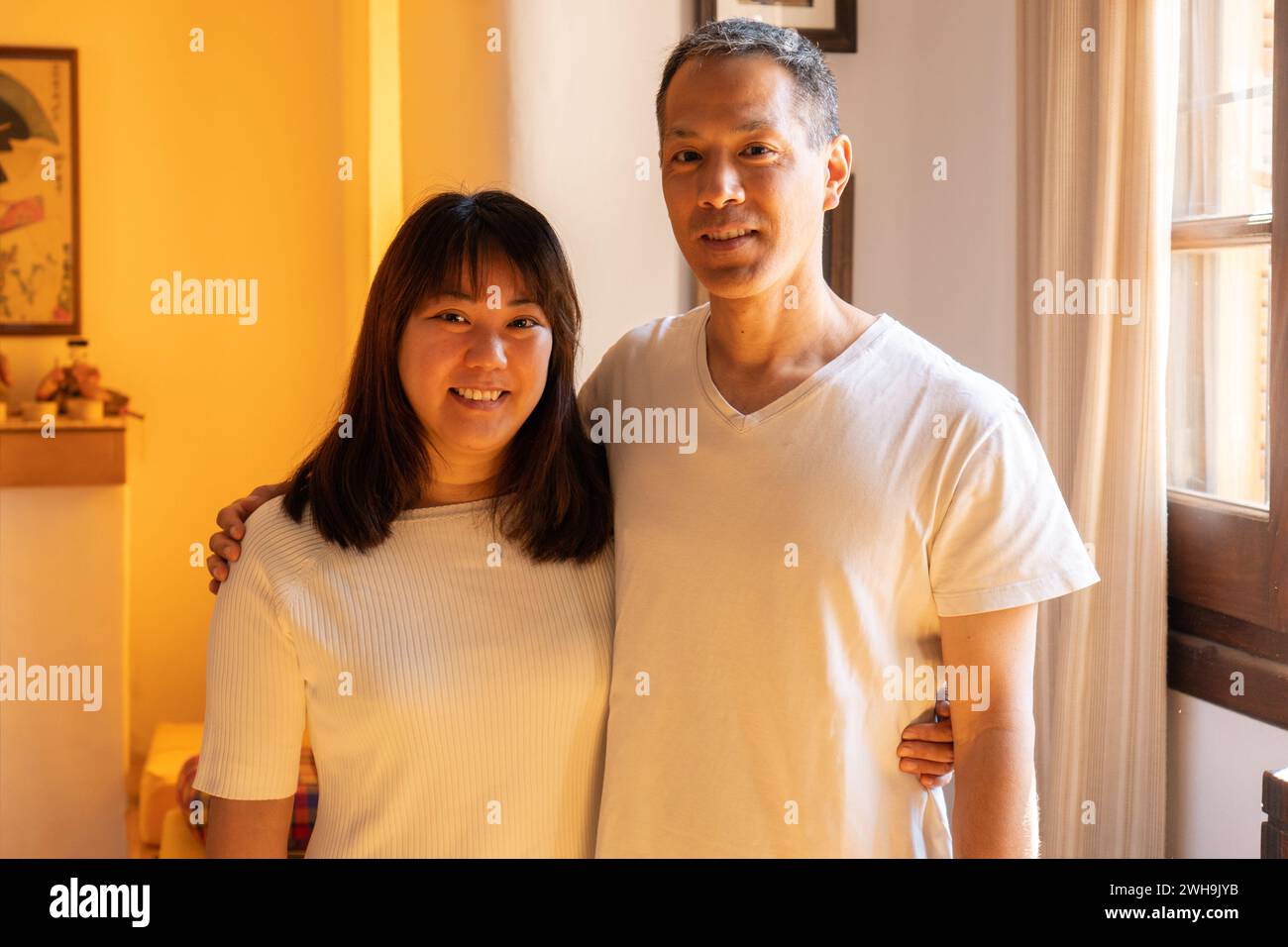 Fratelli adulti in piedi che si abbracciano l'un l'altro di etnia giapponese, sorridono e guardano la telecamera. Verticale orizzontale in interni con luce calda. Foto Stock