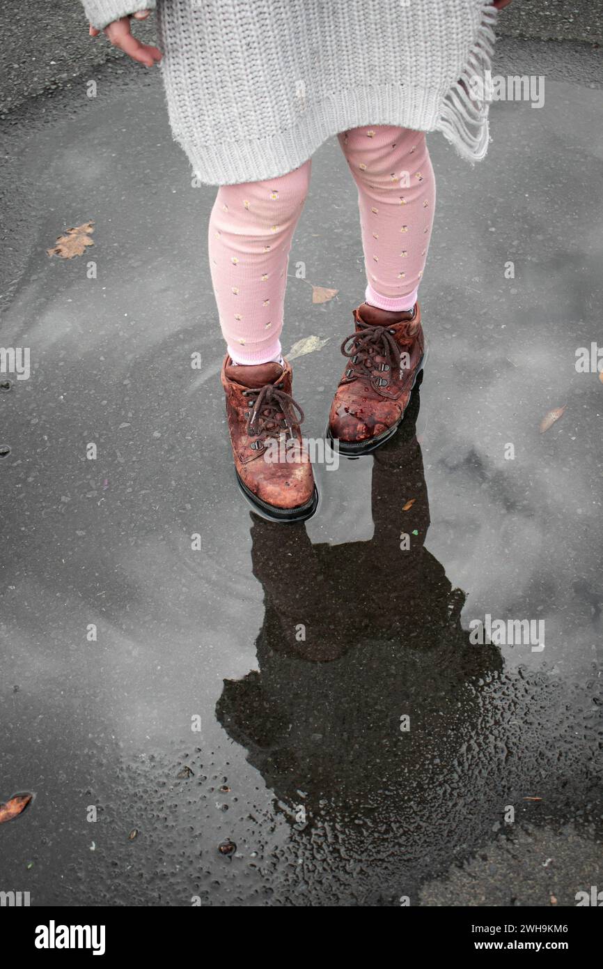 Ragazza con collant rosa, maglione grigio e scarpe marroni che si riflettono in un sottofondo di pioggia con increspature sulla strada Foto Stock