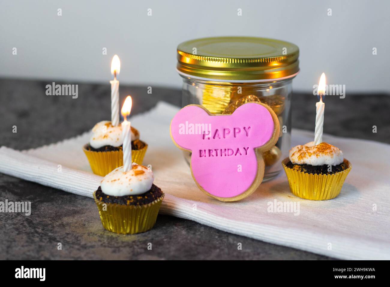 Il biscotto rosa per il topo di buon compleanno sormontato su un vaso color oro con cupcake al cioccolato con glassa di marshmallow bianco e candele di compleanno bianche Foto Stock