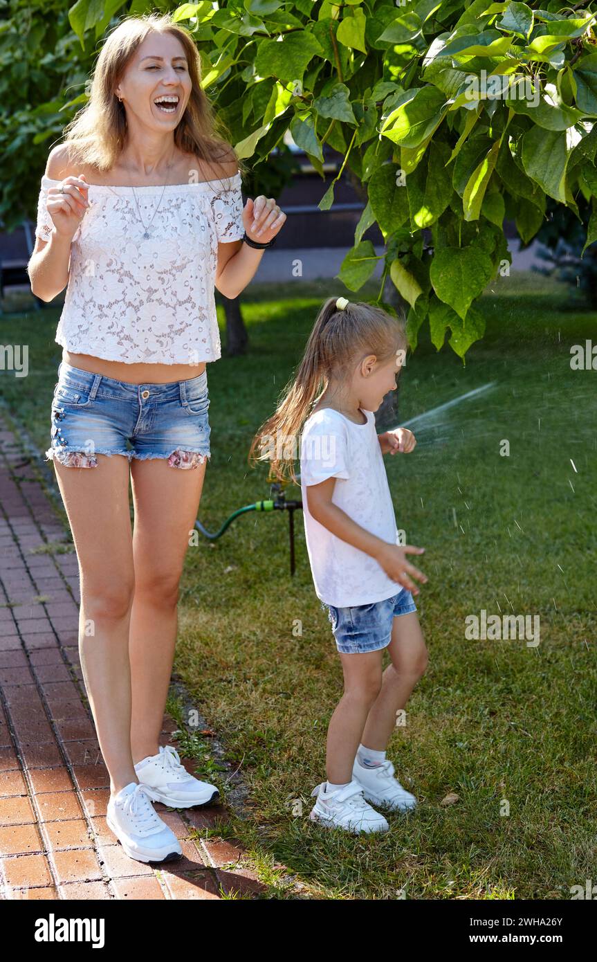 Madre con figlia che cammina sotto l'acqua dall'irrigazione sprinkler nel parco cittadino estivo. Concetto di infanzia, tempo libero e persone - Happy fami Foto Stock