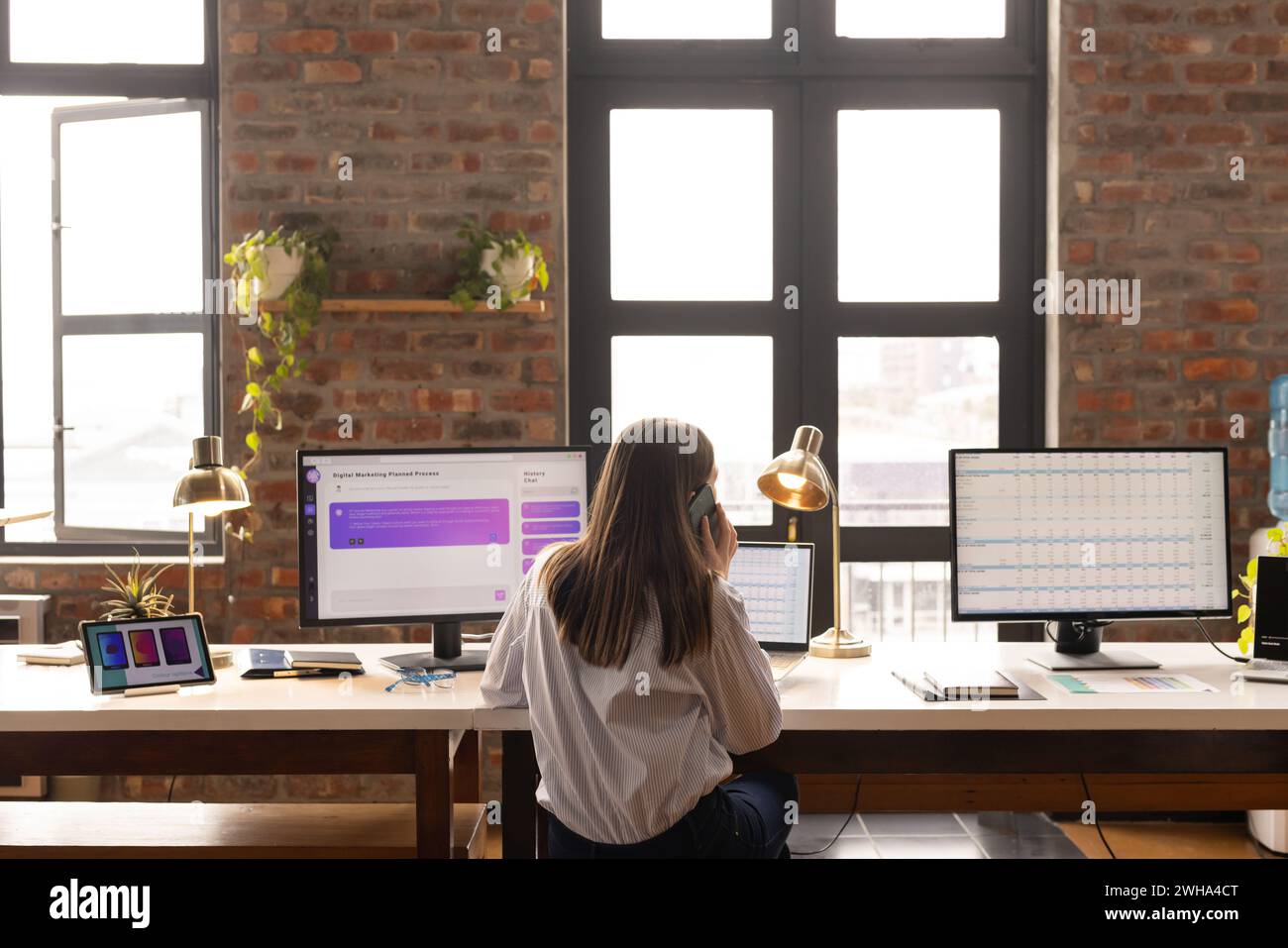 Una giovane donna caucasica lavora su una scrivania con due monitor in un ufficio aziendale informale Foto Stock