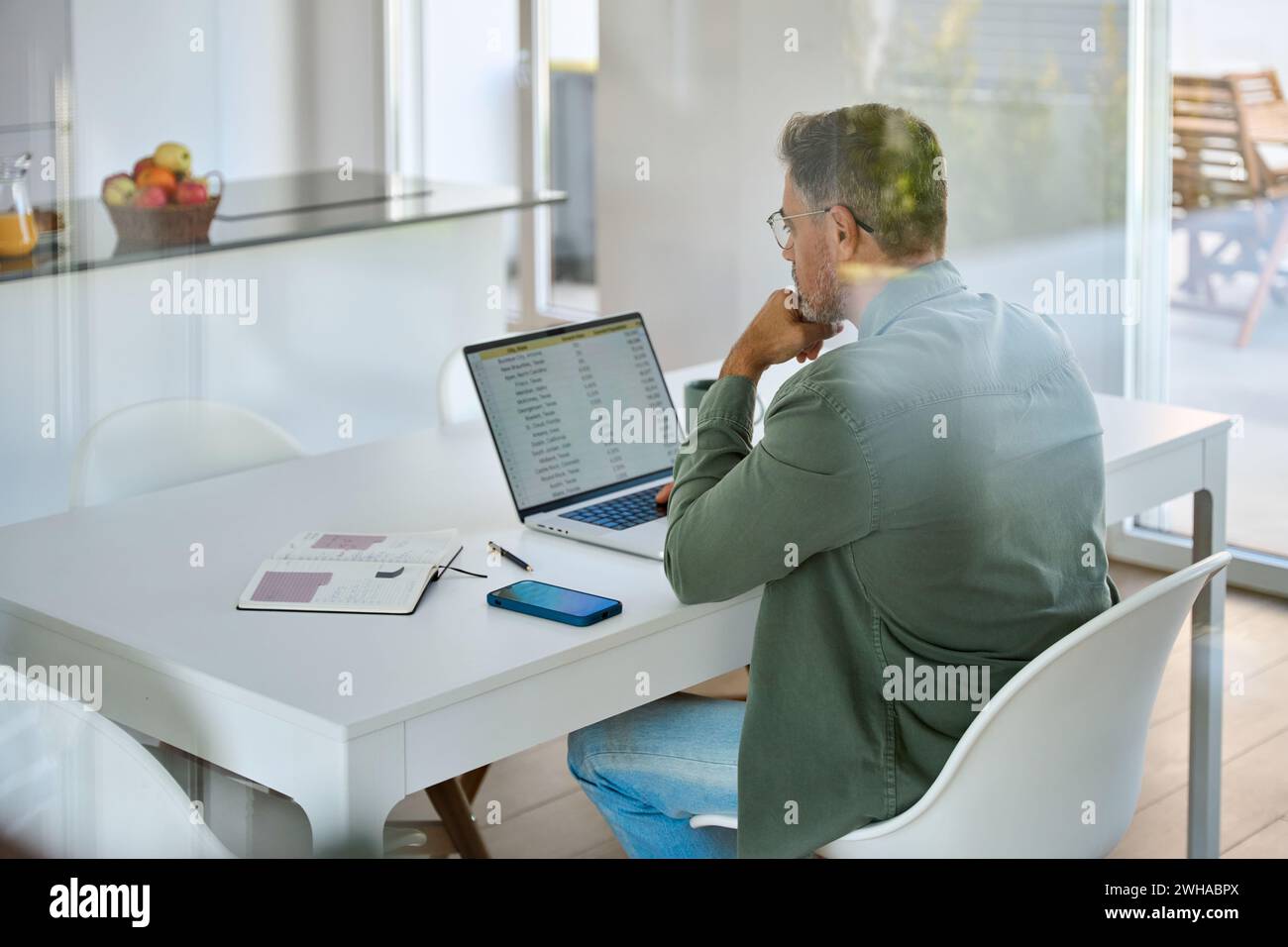 Uomo maturo che guarda il computer seduto al tavolo della cucina ibrido che lavora a casa. Foto Stock
