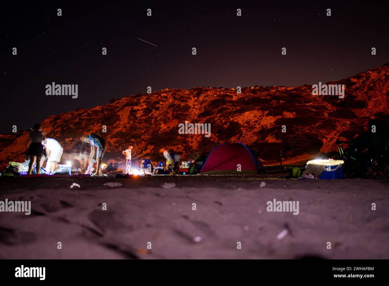 Campeggiatori sotto cieli stellati con tende e luci da campeggio, annidati dal mare e da una montagna rocciosa, creando un ambiente di campeggio costiero sereno Foto Stock
