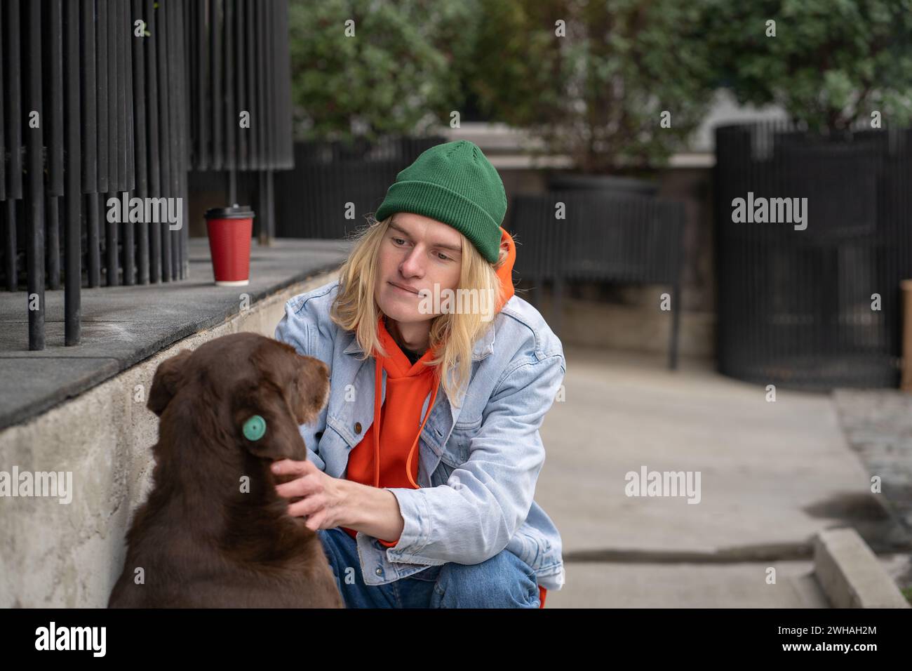 Un ragazzo elegante e cordiale, con un abbigliamento alla moda, seduto su stregoni, accarezzando il cane di strada con amore Foto Stock