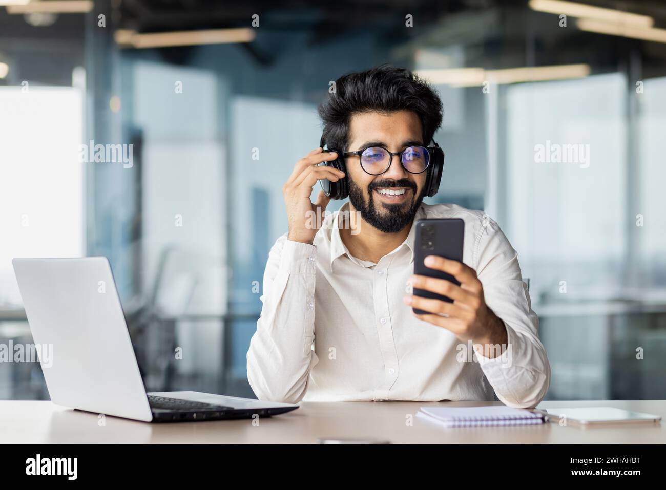 Un uomo con gli occhiali si siede a una scrivania utilizzando un computer portatile e un telefono cellulare, mentre indossa cuffie e occhiali. Ha un sorriso in faccia. Foto Stock