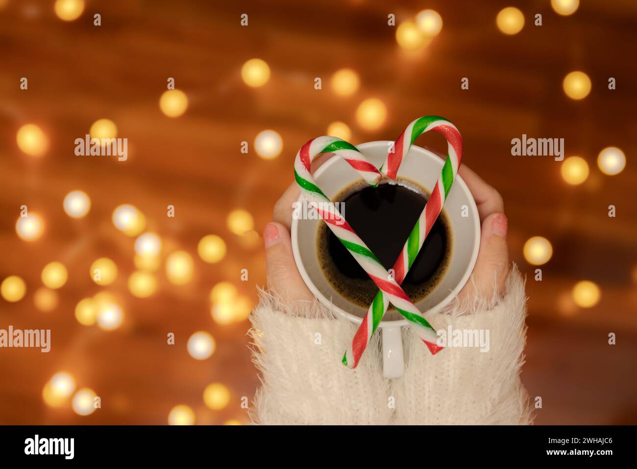 Donna mano con maglione che regge una tazza di caffè con canne di caramelle a forma di cuore sulla parte superiore, decora con luce bokeh sfocata per la festa di San Valentino Foto Stock