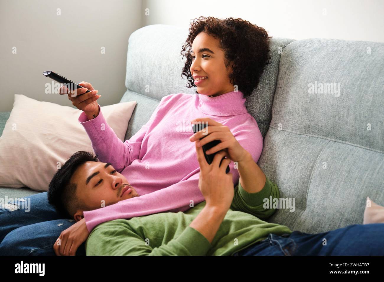 Coppia multietnica che guarda la TV e usa il telefono insieme sul divano. Foto Stock