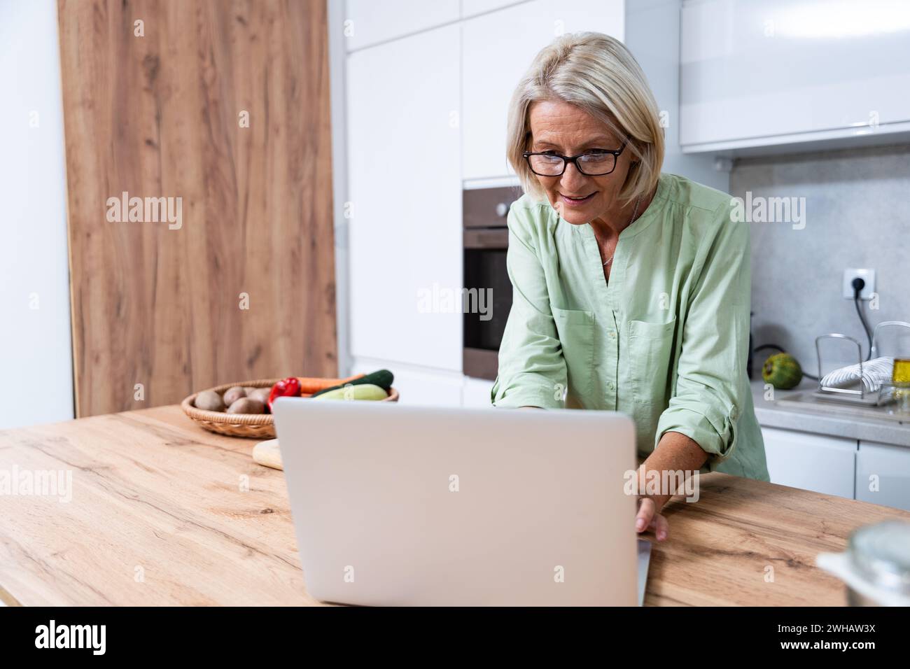 Senior donna sopravvissuto cancro lettura ricette in cucina da computer portatile cucinare sano verdure cibo vegan per una migliore salute e corpo rigenerato Foto Stock