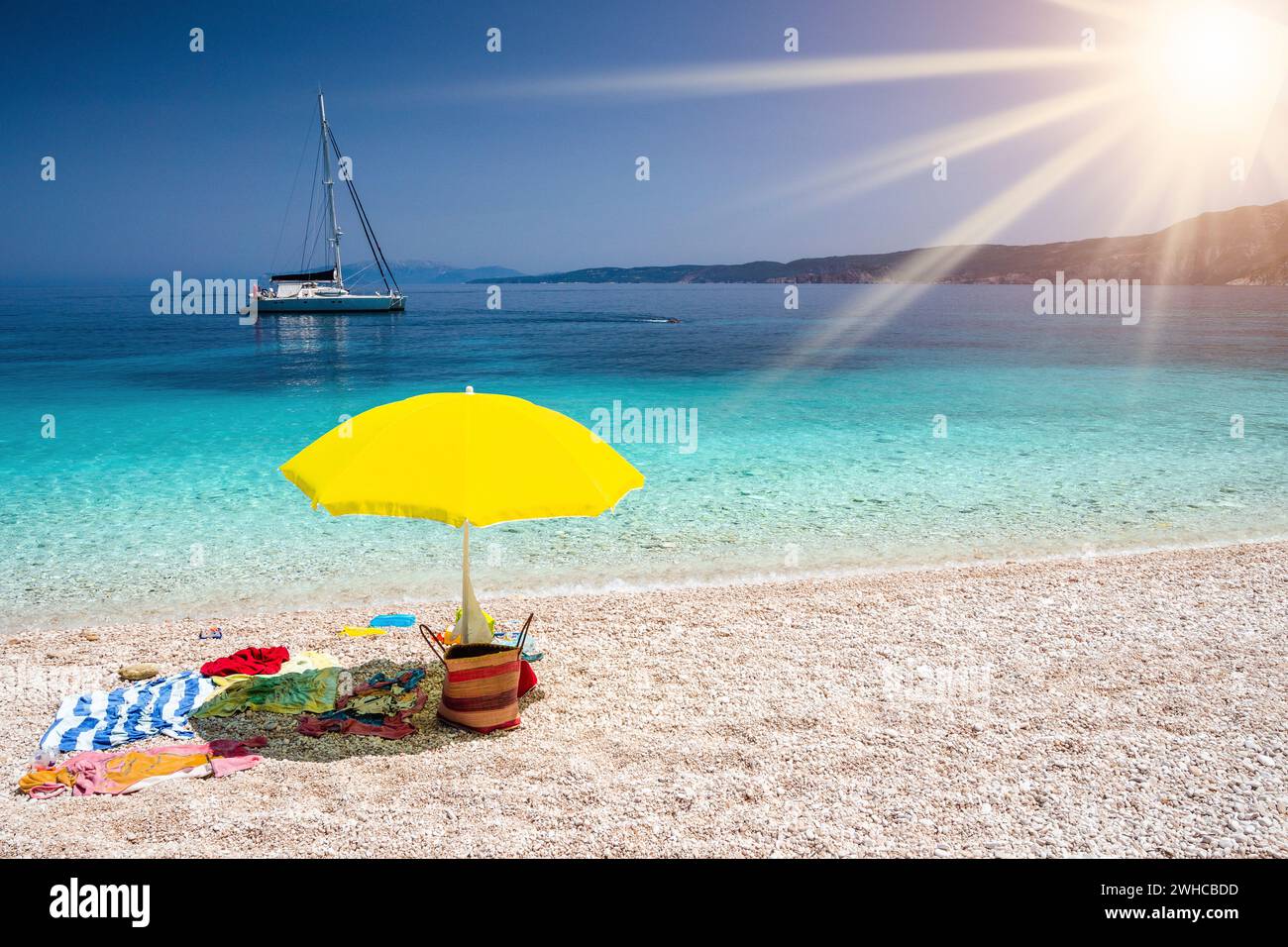 Concetto estivo con accessori in spiaggia. Ombrello giallo su pittoresco sacchetto di paglia di ciottoli, asciugamano, isola di Cefalonia, Grecia. Concetto di vacanza relax in viaggio. Foto Stock