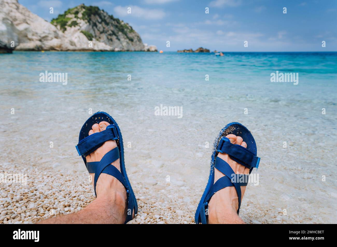I giovani piedi maschili indossano sandali infradito blu che prendono il sole sulla spiaggia di ciottoli di fronte all'acqua blu del mare e le rocce sullo sfondo all'orizzonte. Foto Stock