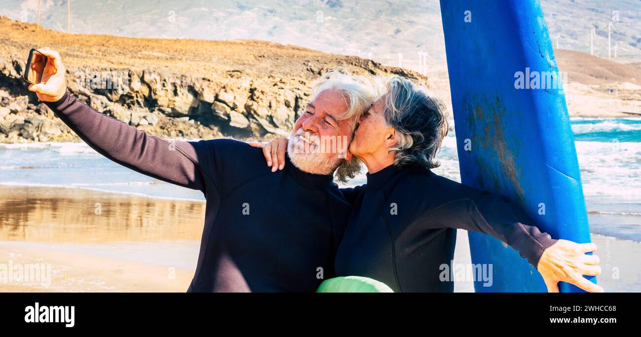 coppia di due anziani carini e pensionati che fanno esercizio e. prendere un selfie con i loro tavole da surf e muta - donna baciare il marito Foto Stock