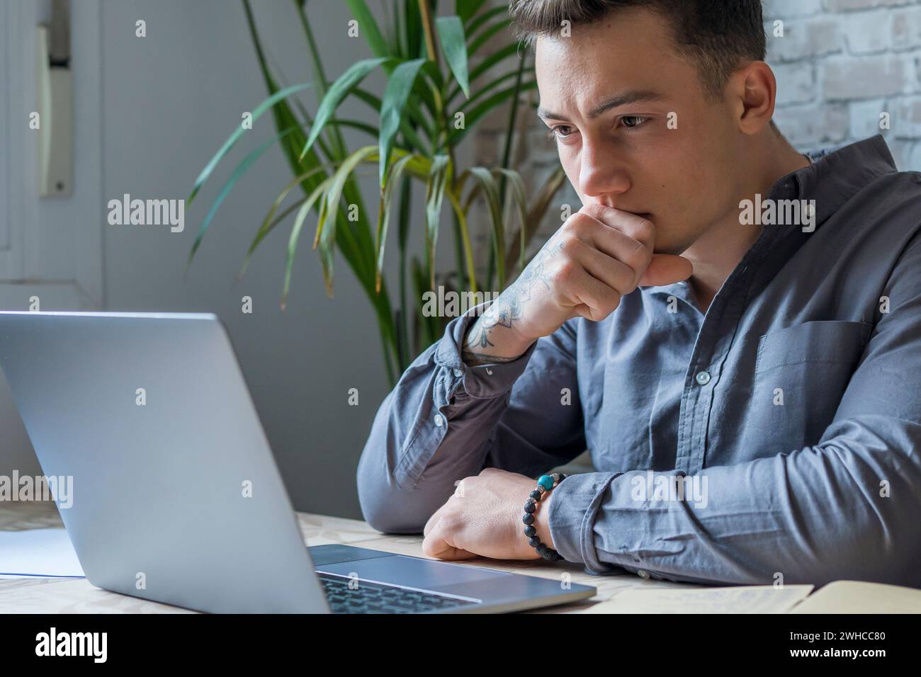 Uomo d'affari premuroso che tocca il mento, riflette idee o strategie, siede alla scrivania di legno con un computer portatile, lavora come libero professionista su progetti online, stud Foto Stock