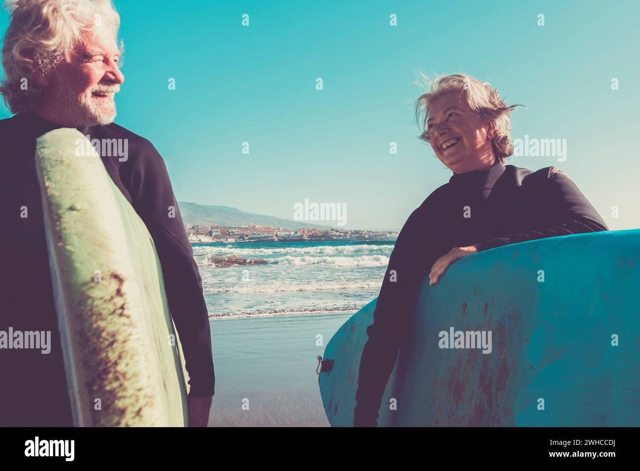 coppia di anziani in spiaggia con mute nere che tengono un tavolo da surf pronto a navigare in spiaggia - persone attive mature e pensionati che fanno felice attività insieme in le loro vacanze o tempo libero Foto Stock