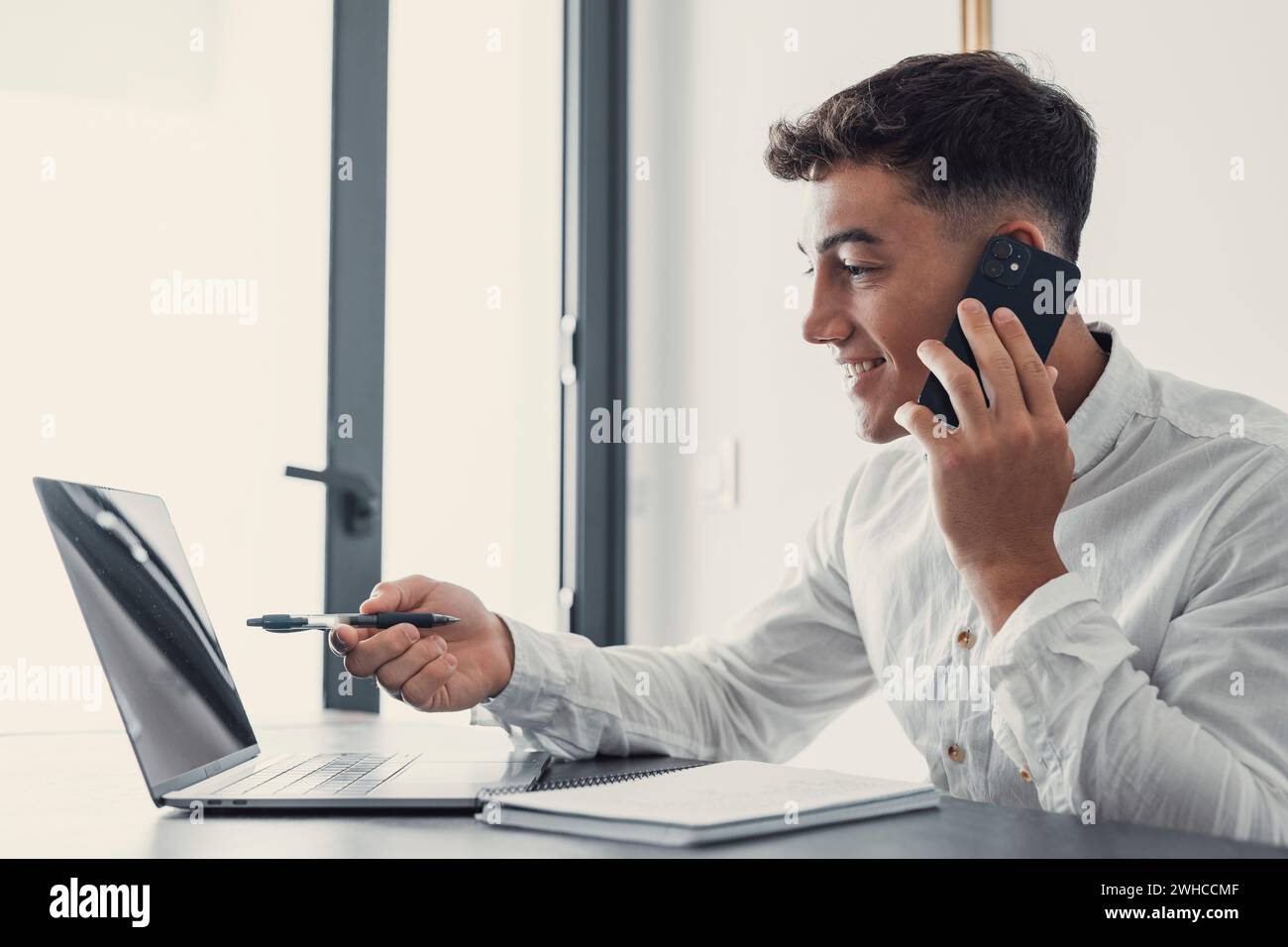 Un uomo d'affari sorridente che indossa occhiali che parlano al telefono, seduto alla scrivania con un computer portatile, un manager cordiale che si occupa della consulenza telefonica con i clienti, un uomo felice che chiacchiera con gli amici distratto dal lavoro Foto Stock