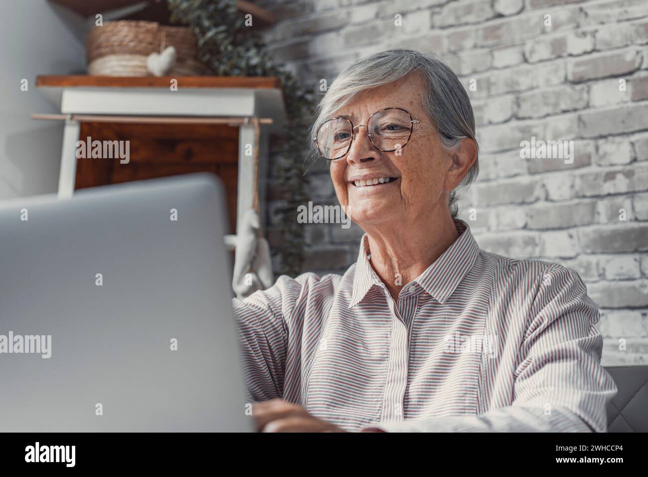Lavoratore remoto anziano. Una donna anziana concentrata negli occhiali lavora su un computer portatile dall'ufficio domestico leggendo i documenti elettronici delle e-mail. Lavoratrice freelance anziana siede al tavolo della cucina per pc scrivendo online Foto Stock
