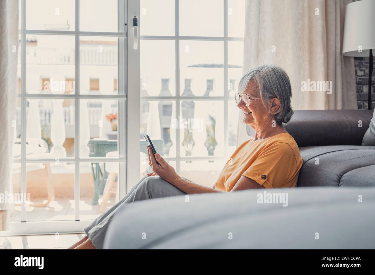 Sorridente donna caucasica di mezza età seduto accanto al divano in soggiorno a navigare su Internet wireless al telefono, felice donna anziana moderna rilassarsi a terra a casa utilizzando il dispositivo smartphone, concetto di tecnologia per anziani Foto Stock