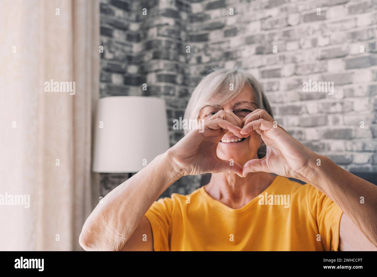 Donna anziana seduto in salotto dita connesse che mostrano il simbolo del cuore da vicino, anziani trattamento di prevenzione delle malattie cardiovascolari, controllo di salute, vitamine cardio, segno di gentilezza e carità Foto Stock