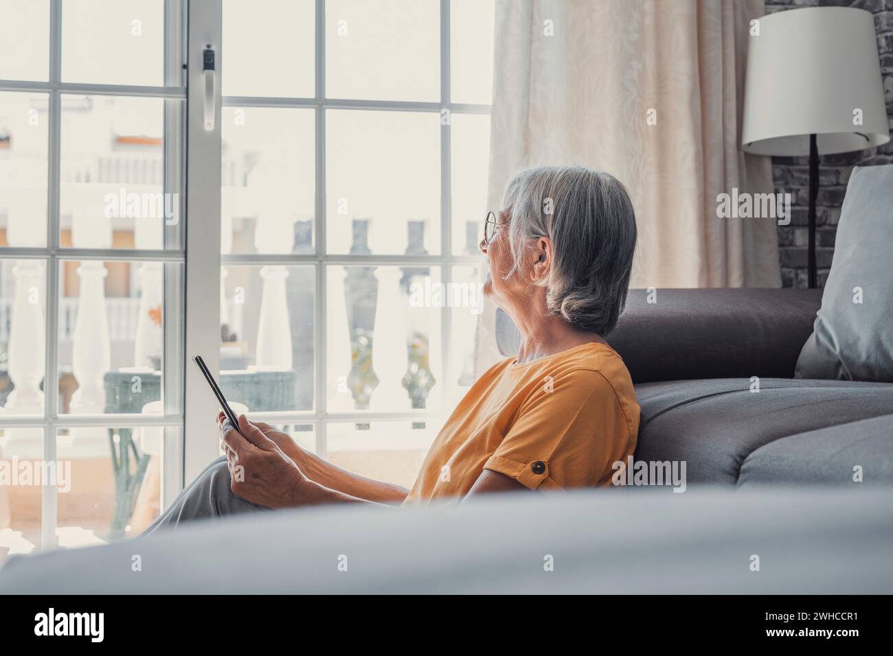 Sorridente donna caucasica di mezza età seduto sul divano in soggiorno a navigare su Internet wireless su tablet, felice donna anziana moderna rilassarsi a terra a ho Foto Stock