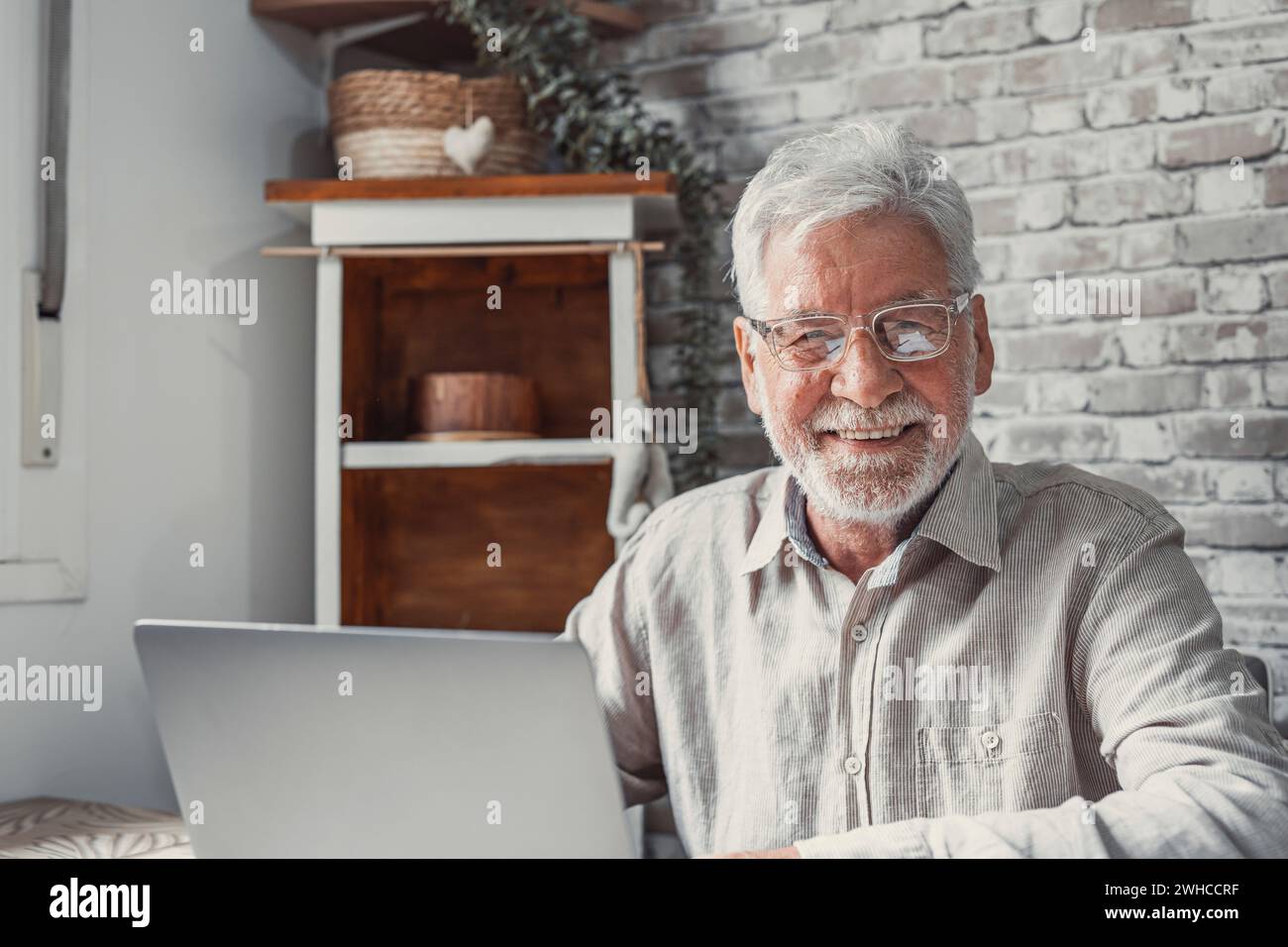 Lavoratore remoto anziano. Un uomo anziano concentrato negli occhiali lavora su un laptop dall'ufficio domestico guardando la fotocamera. Un dipendente di vecchiaia freelance siede al tavolo della scrivania per pc scrivendo un report online Foto Stock
