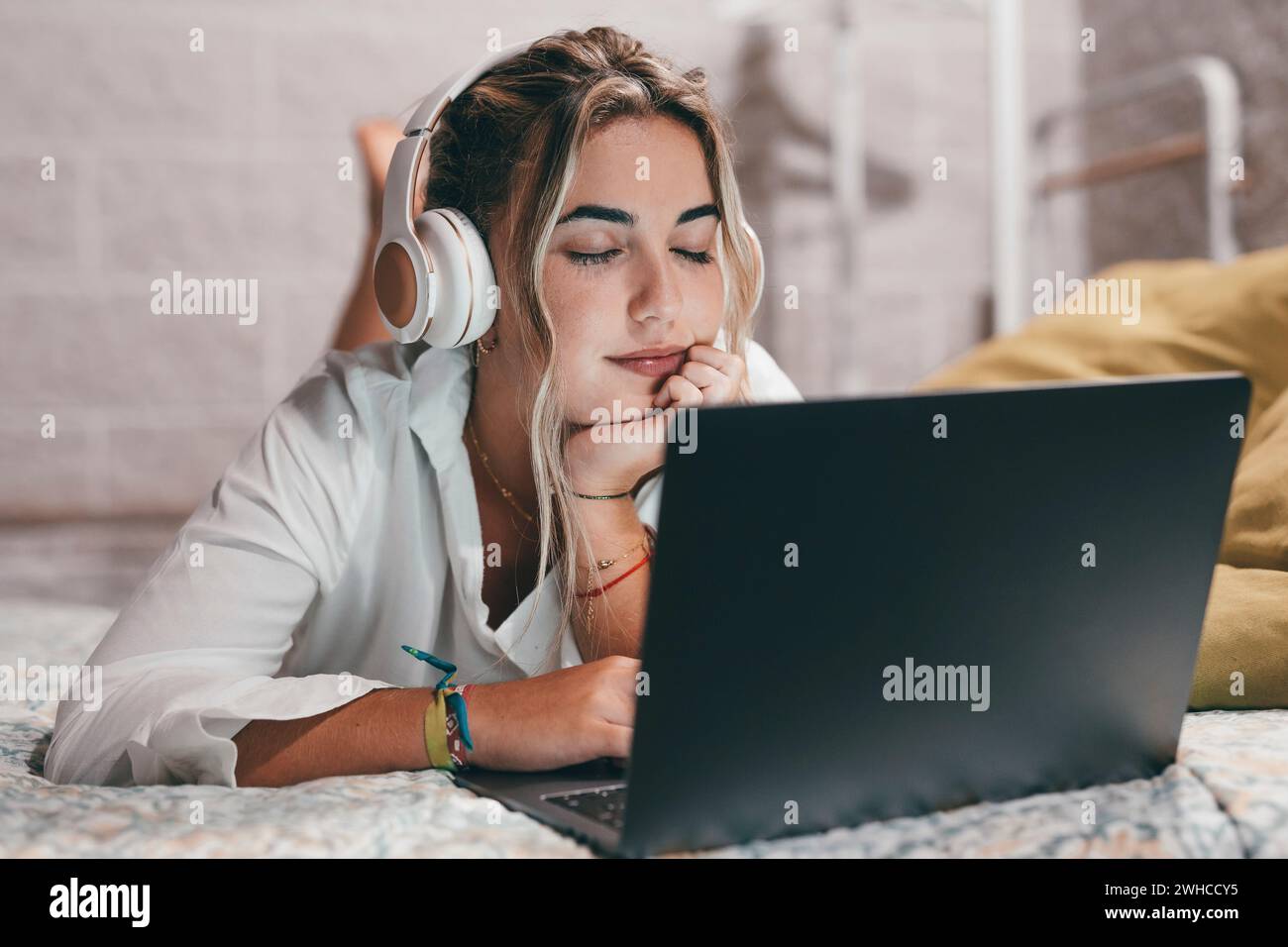Primo piano di una giovane donna felice che usa il laptop in casa sdraiato sul letto e indossa le cuffie per ascoltare musica. Adolescente millenario che si rilassa ascoltando canzoni a occhi chiusi Foto Stock