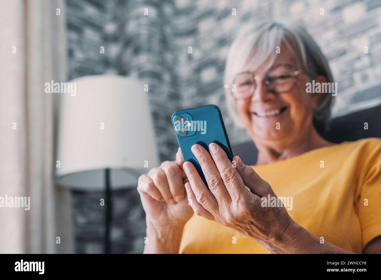 Sorridente donna caucasica di mezza età seduto accanto al divano in soggiorno a navigare su Internet wireless al telefono, felice donna anziana moderna rilassarsi a terra a casa utilizzando il dispositivo smartphone, concetto di tecnologia per anziani Foto Stock