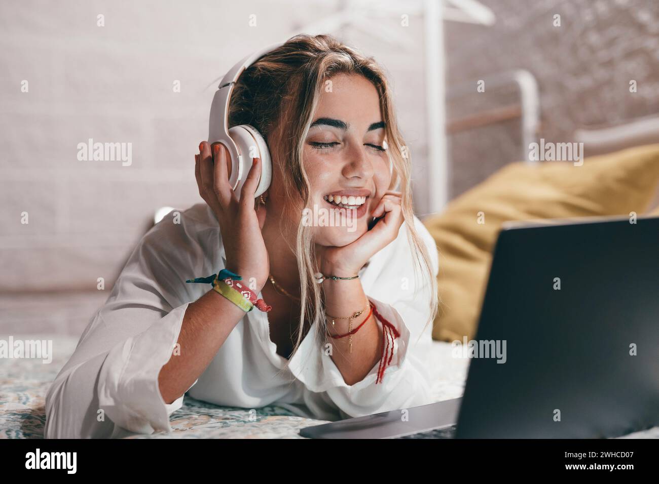 Primo piano di una giovane donna felice che usa il laptop in casa sdraiato sul letto e indossa le cuffie per ascoltare musica. Adolescente millenario che si rilassa ascoltando canzoni a occhi chiusi Foto Stock