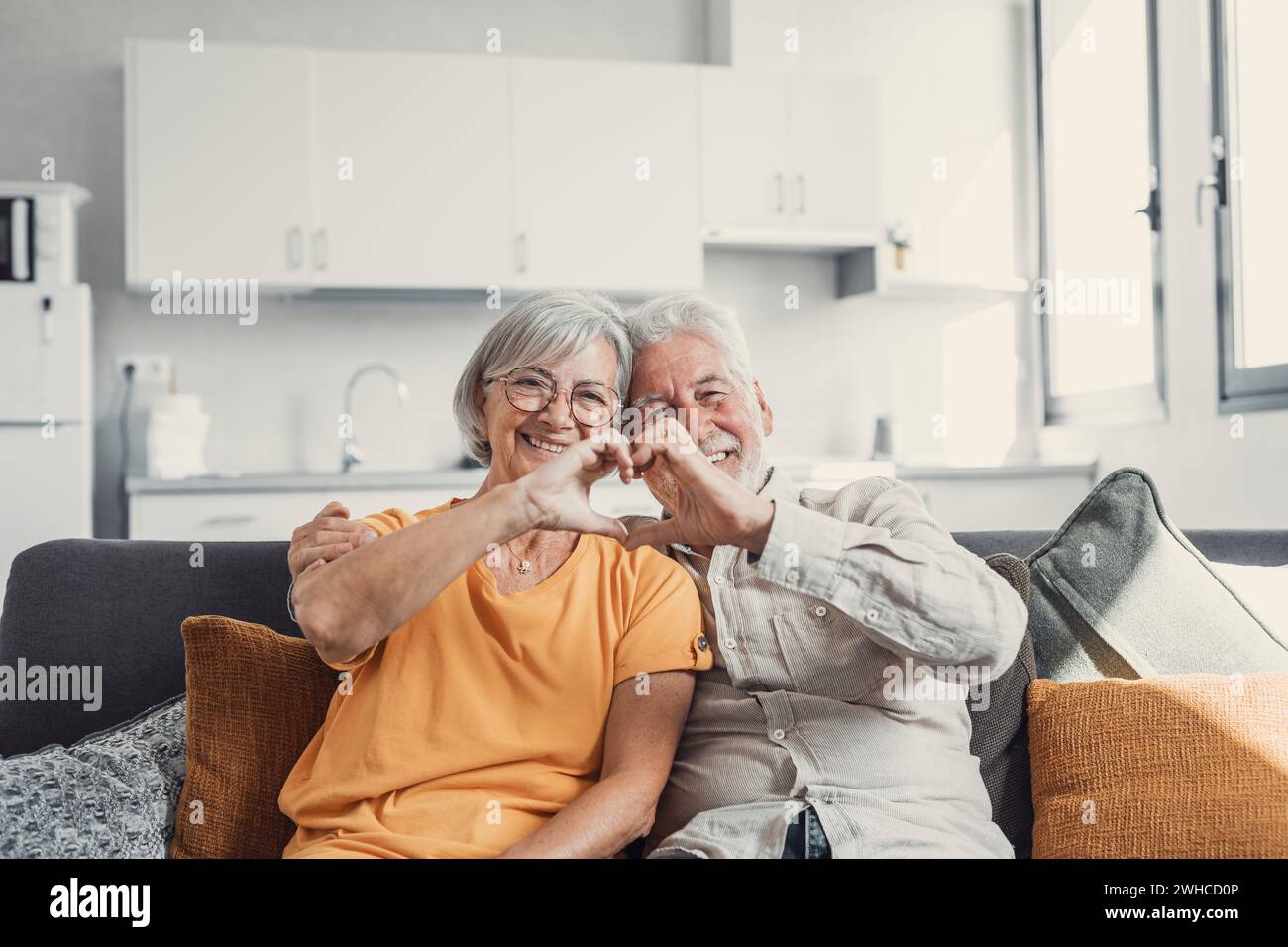 Primo piano ritratto felice sincero anziano di mezza età pensionato coppia di famiglia facendo gesto cardiaco con le dita, mostrando l'amore o dimostrando sentimenti sinceri insieme in interni, guardando la fotocamera. Foto Stock