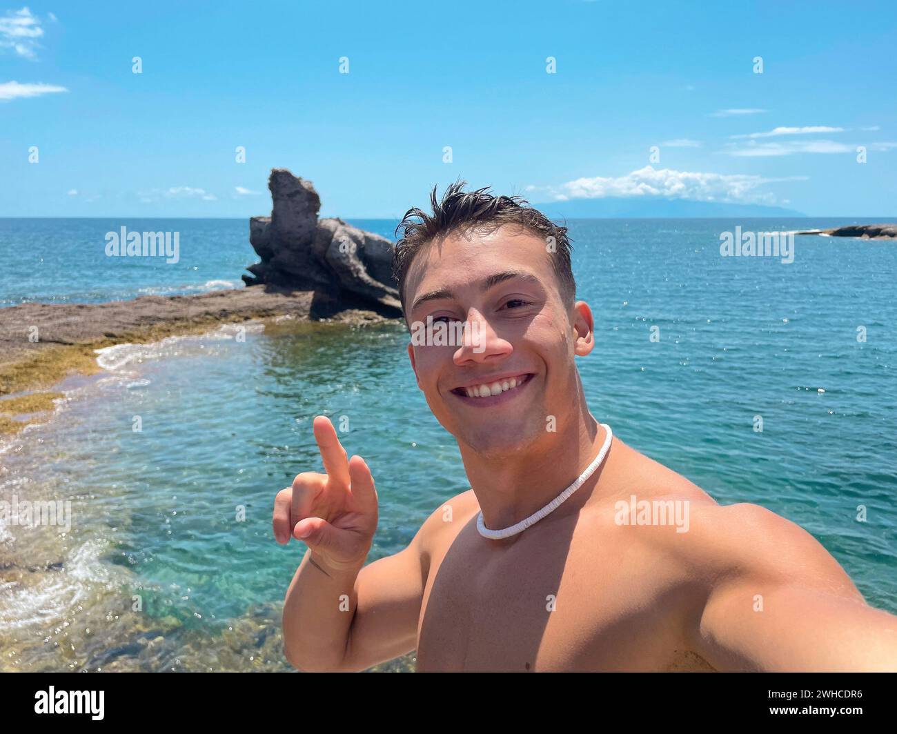 Un giovane attraente e felice in spiaggia sorride e guarda la fotocamera che tiene e scatta un selfie con il suo telefono. Tempo estivo in vacanza divertendosi e divertendosi Foto Stock