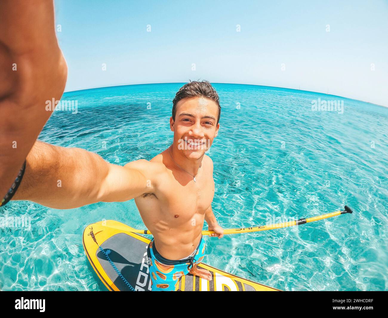 Un uomo caucasico felice che si diverte e si diverte in vacanza all'aperto sulla spiaggia, facendo paddle surf in acqua. Un ragazzo attraente che si sente libero di viaggiare. Foto Stock