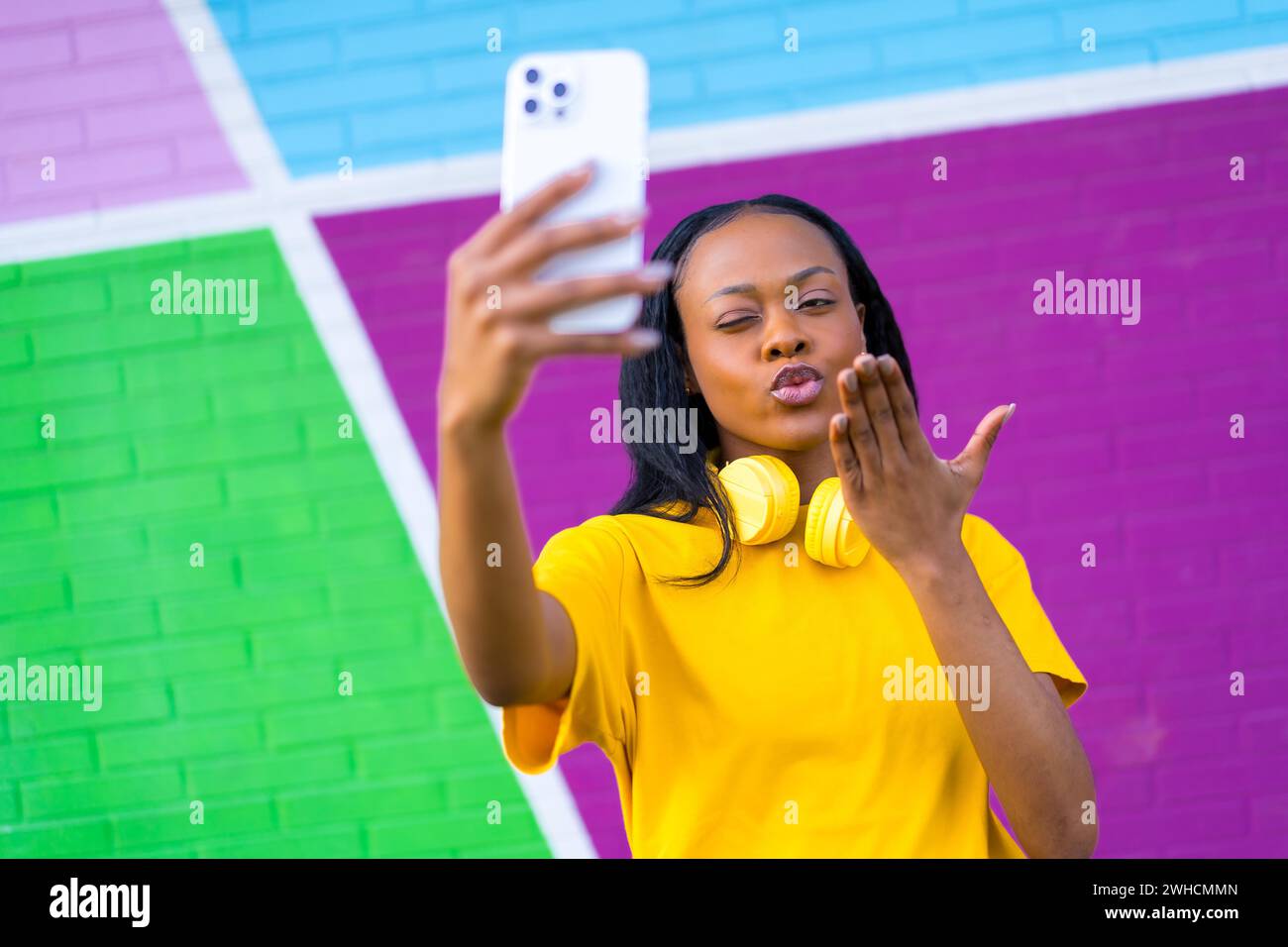 Donna africana che soffia un bacio mentre scatta un selfie contro una colorata parete urbana all'aperto Foto Stock