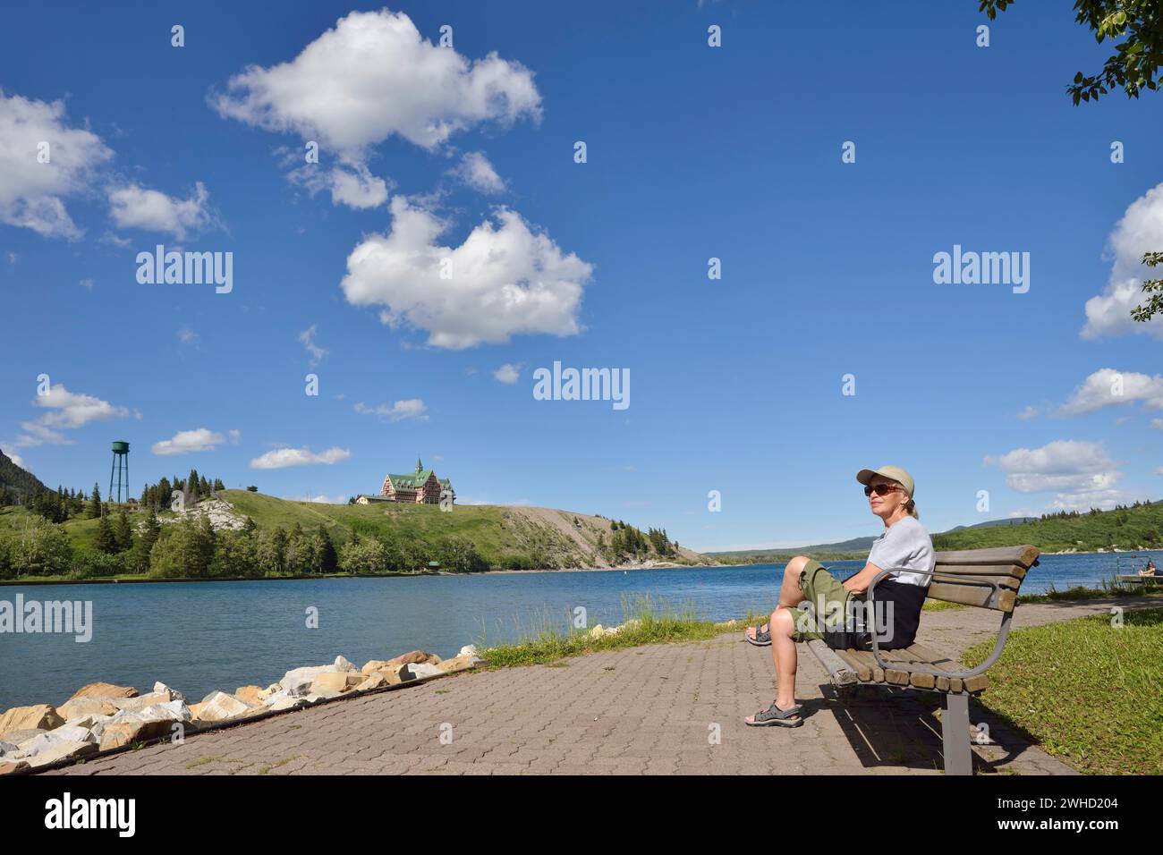 Donna seduta su una panchina sulla riva del lago Upper Waterton con vista sul Prince of Wales Hotel, Waterton Lakes National Park, Alberta, Canada Foto Stock