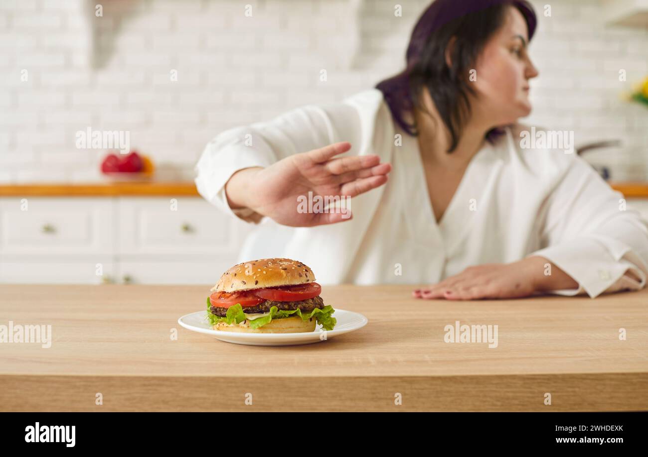 La donna grassa inizia a mangiare, dice DI NO al cibo malsano e spinge via il piatto con l'hamburger Foto Stock