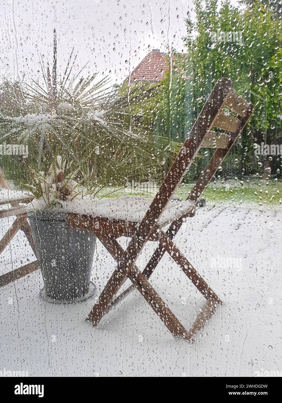 Guardando fuori attraverso le gocce di pioggia sulla finestra le piante verdi e il fenomeno meteorologico della neve a maggio a Berlino a causa dei cambiamenti climatici Foto Stock