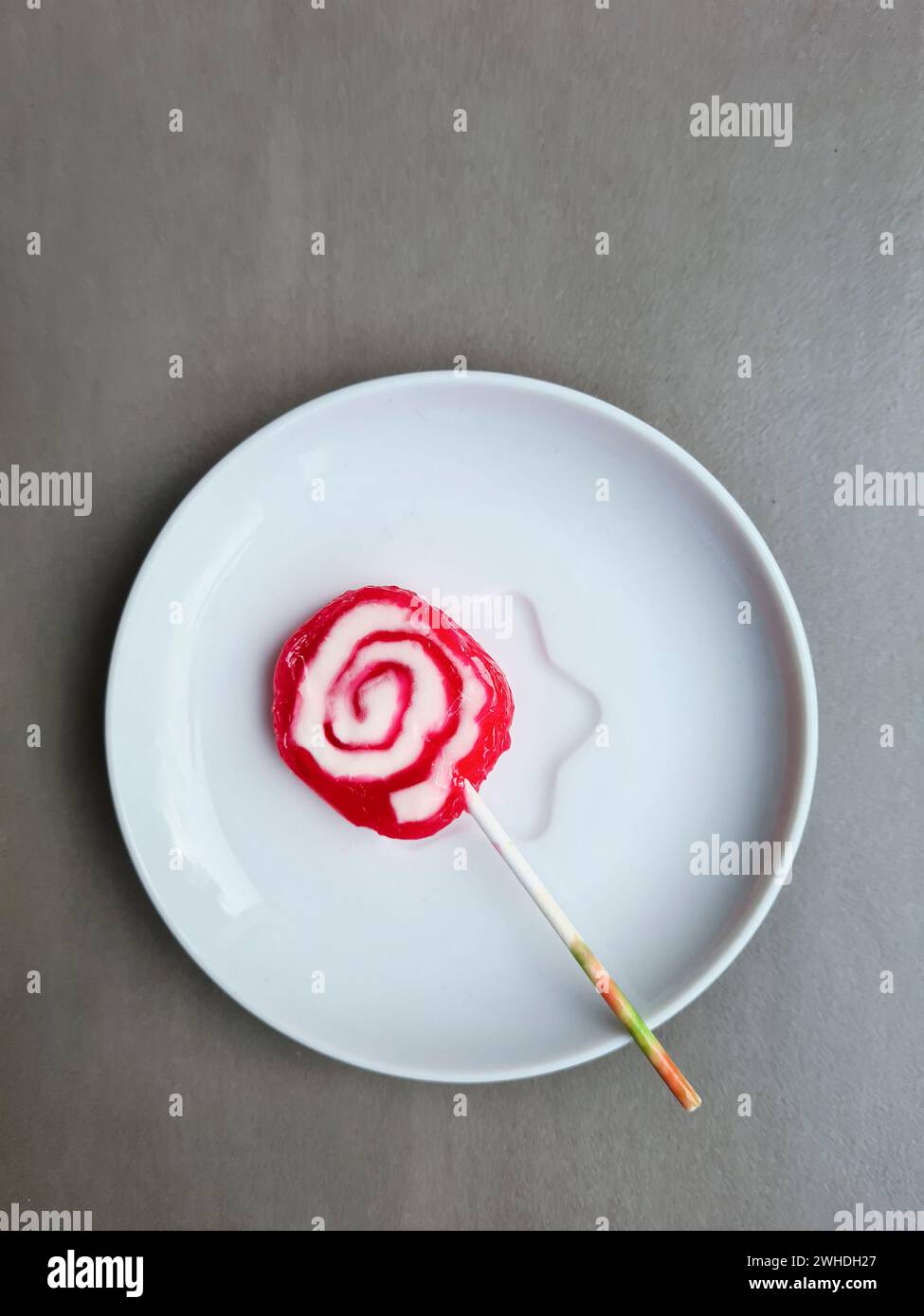 Un lecca lecca rosso e bianco con un bastoncino giace su un piatto su uno sfondo grigio chiaro Foto Stock