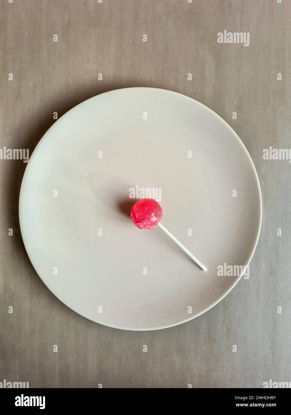 Un lecca lecca rosso con un bastoncino giace su un piatto su uno sfondo grigio chiaro Foto Stock