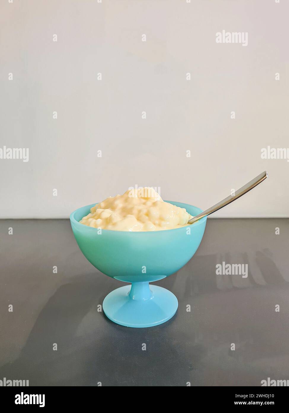 Dessert, budino di riso con cucchiaio in una ciotola di vetro su un piatto da cucina grigio su uno sfondo bianco alla luce del giorno Foto Stock
