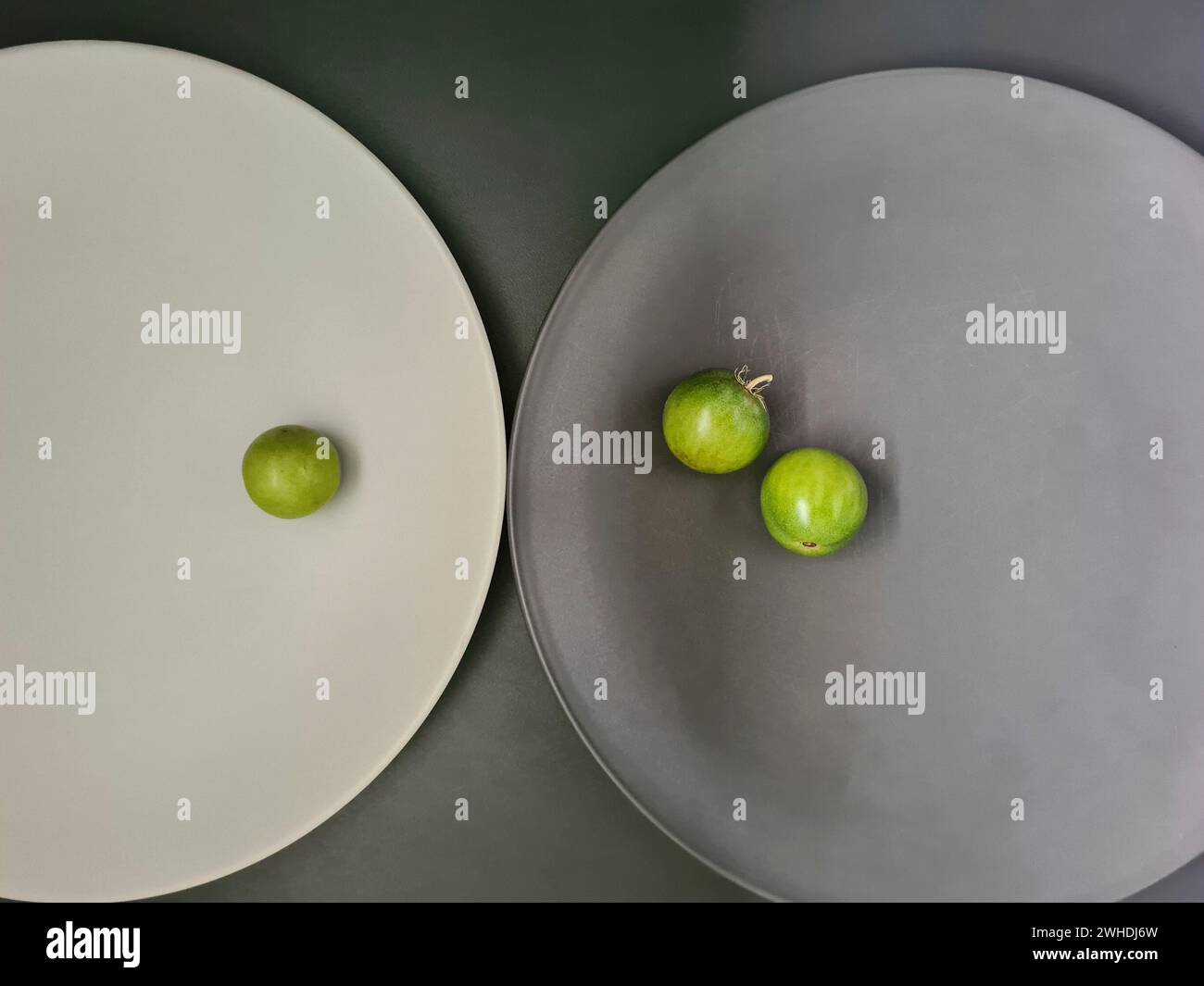 un piatto bianco con 1 pomodoro verde e un piatto grigio con 2 pomodori piccoli su sfondo grigio sul tema dei contrasti e del minimalismo Foto Stock