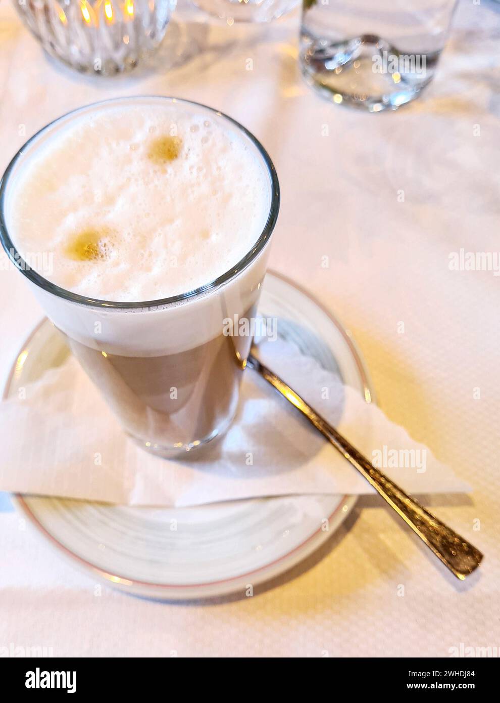Caffè con schiuma di latte in un bicchiere su un piattino e cucchiaio su un supporto per tovagliolo su una tovaglia bianca Foto Stock