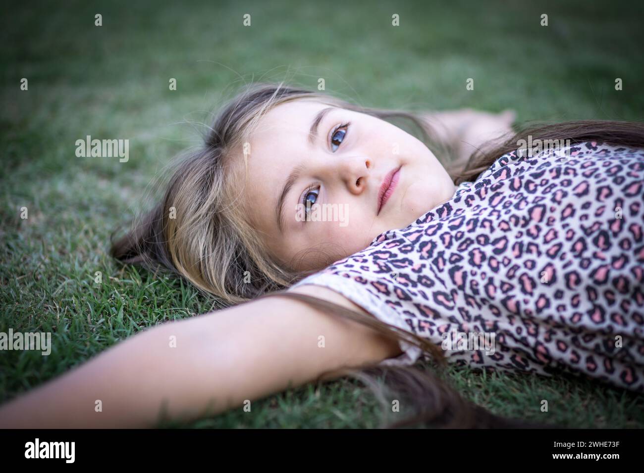 Hermosa muchacha rubia con gracia mientras disfruta de la naturaleza en un escenario al aire libre lleno de verdor y luz y recostada en el césped Foto Stock