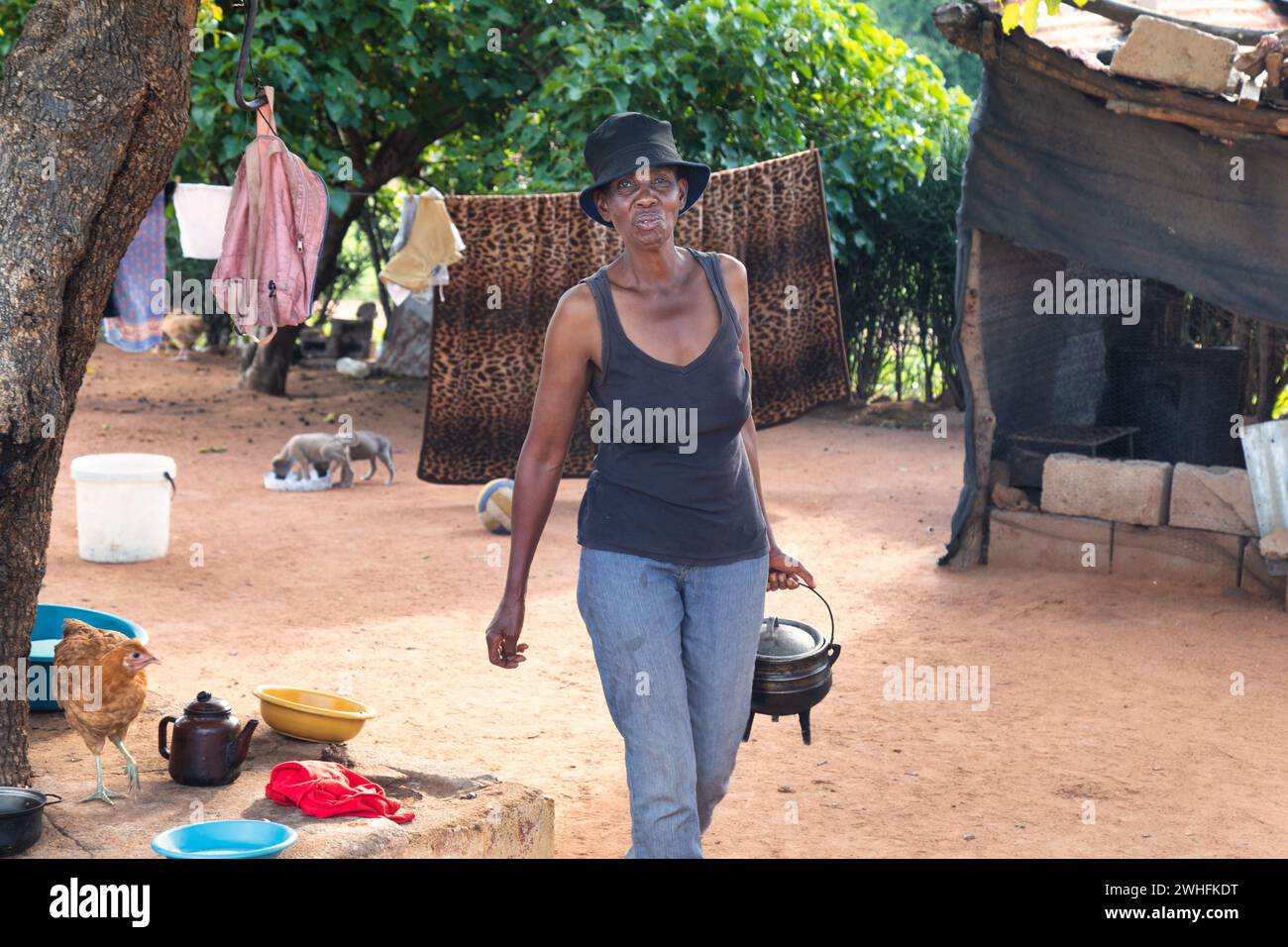 donna africana del villaggio che cammina nel cortile con una pentola a tre zampe in mano, di fronte alla cucina all'aperto Foto Stock