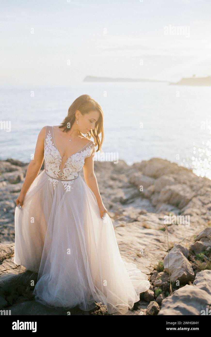 La sposa con un abito soffice bianco si erge su una spiaggia rocciosa che tiene l'orlo con le mani Foto Stock