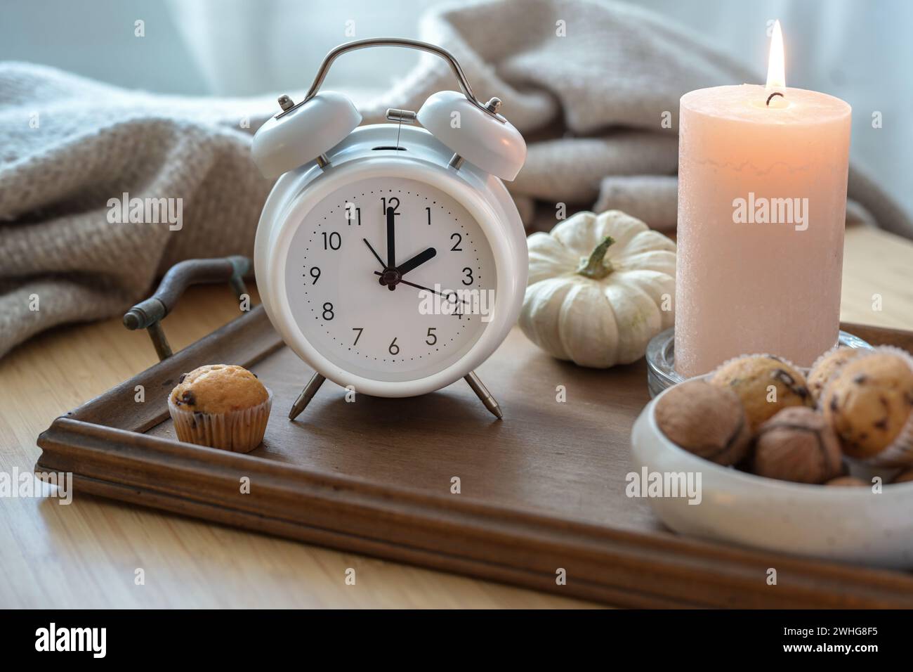 Sveglia vintage bianca, candela, decorazioni autunnali e dolci su un comodino con coperta naturale di lana, concetto di ripiegamento Foto Stock