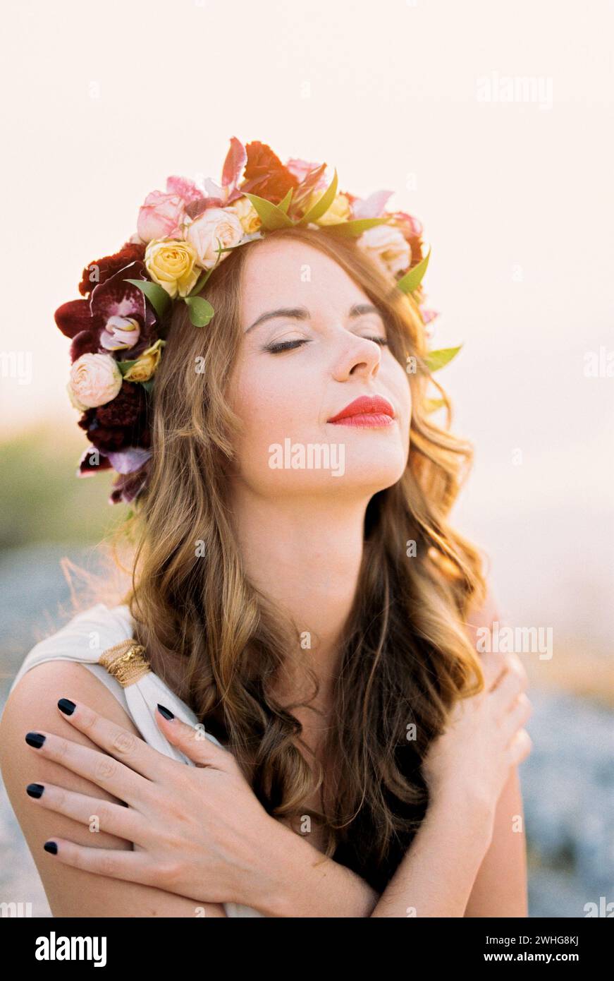 Sposa sorridente in una corona di fiori che si abbraccia alle spalle, sollevando la testa al sole Foto Stock