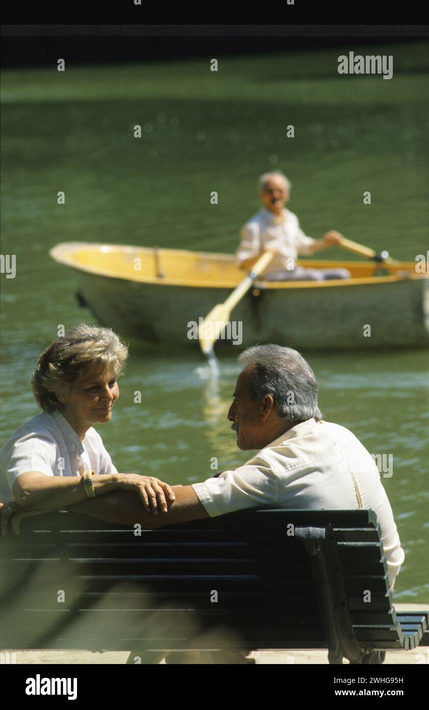 una coppia di anziani si siede romantica su un'ench che si guarda sullo sfondo del lago con gente che canta Foto Stock