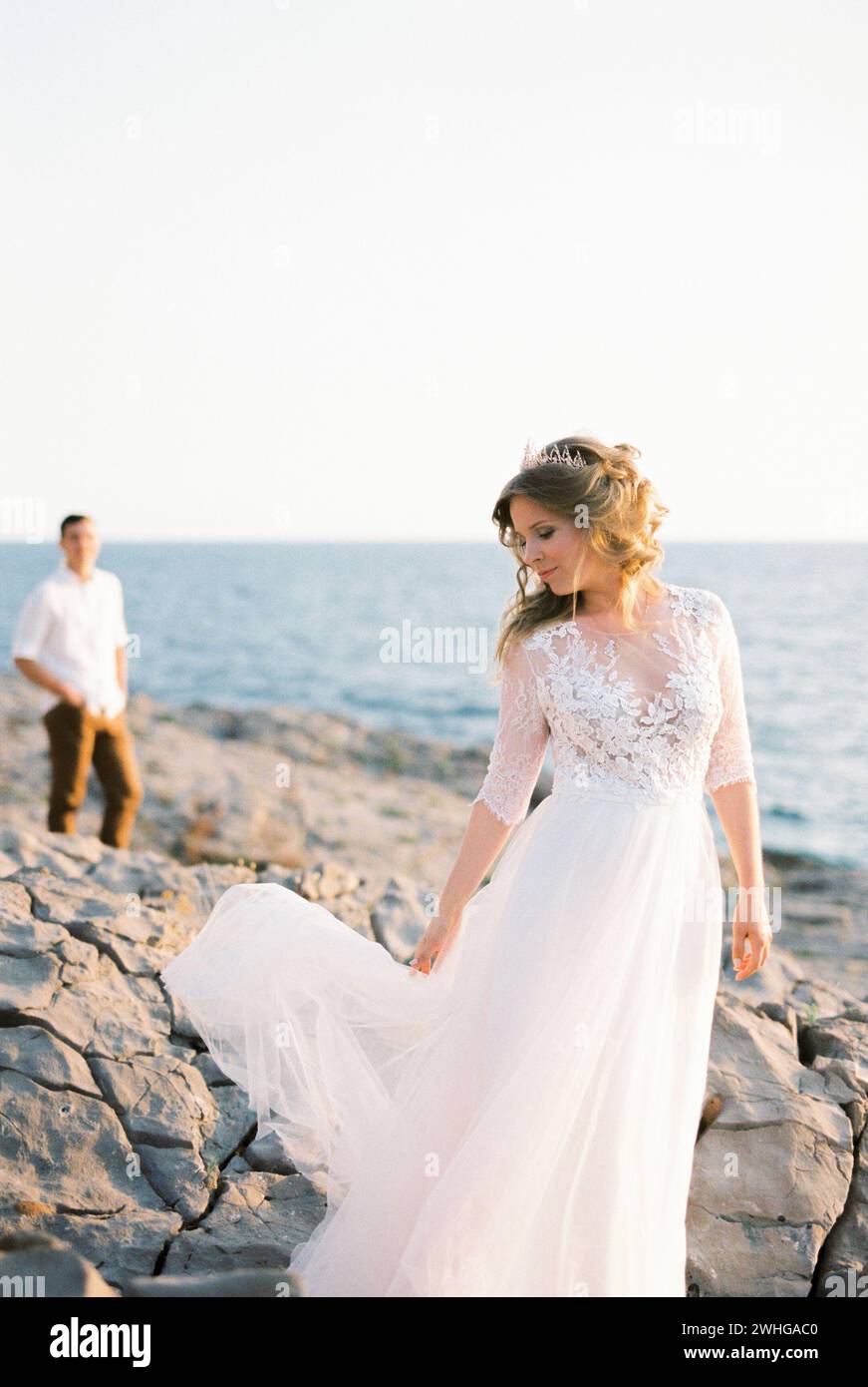 Sposa solleva l'orlo del suo vestito sulle rocce vicino al mare sullo sfondo dello sposo Foto Stock